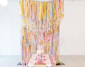 May include: A rectangular white table with pink chairs and a gold sequin runner, set with pink plates and cups. Behind the table hangs a large backdrop made of long, vertical strips of pastel pink, blue, yellow, and metallic gold streamers.