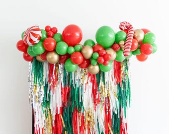 May include: A festive balloon garland featuring red, green, and gold balloons, accented with a red and white striped peppermint candy balloon and a candy cane balloon, hanging above a backdrop of red, green, white, and gold metallic fringe.