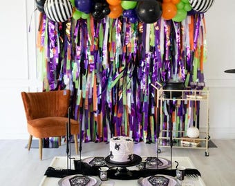 May include: A Halloween party setup featuring a balloon arch in orange, purple, green, and black, with black and white striped balloons. A white table holds a cake with spider web designs, coffin-shaped plates, and black cups. A fringe backdrop hangs behind.