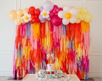 May include: A party backdrop featuring a vibrant fringe curtain in shades of orange, pink, red, and purple, topped with a balloon arch including daisy-shaped clusters. A white table holds a small cake, disco balls, and cups, with floor pillows.
