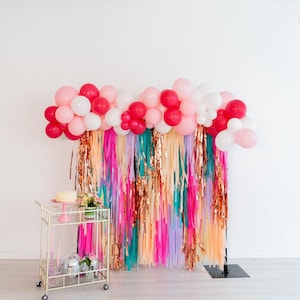 May include: A party backdrop featuring a balloon garland in shades of pink, red, and white, layered over a fringe curtain of metallic copper, teal, lavender, and peach strips. A gold metal bar cart with a cake and disco ball stands to the left.