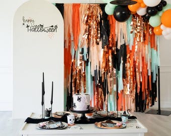 May include: A Halloween party setup featuring a white arched backdrop with the text Happy Halloween, a fringe curtain in orange, black, and mint, a cake with spider decorations, black taper candles, and assorted floor pillows in various colors.