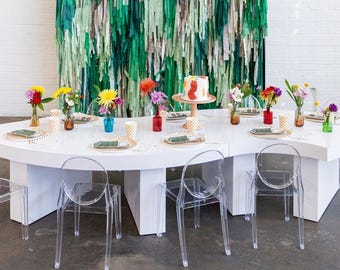 Op de afbeelding: Een grote, witte, gebogen tafel met een glanzende afwerking, omringd door doorzichtige acrylstoelen. De tafel is gedekt met kleine glazen vaasjes met kleurrijke bloemen, papieren bekers met patronen en borden. Een taart staat op een houten standaard.