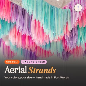 May include: A ceiling installation of hanging paper streamers in shades of bright pink, light purple, and turquoise. The text on the image reads: CUSTOM MADE TO ORDER Aerial Strands Your colors, your size — handmade in Fort Worth.