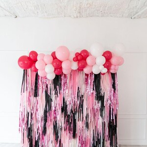 May include: A decorative party backdrop featuring a dense fringe curtain in alternating vertical strips of black, light pink, and metallic hot pink. The top edge is adorned with a cluster of round balloons in shades of white, pink, and red.