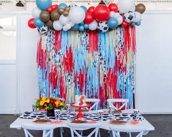 May include: A party setup featuring a white table with a cow-print runner, white chairs, and a backdrop of red, light blue, and cream streamers. A balloon garland in red, blue, white, brown, and cow-print patterns hangs above the table.