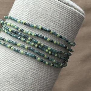 May include: A beaded wrap bracelet with multiple strands. The bracelet features small, colorful beads in shades of teal, green, yellow, and black. The bracelet is wrapped around a neutral-colored, textured surface.