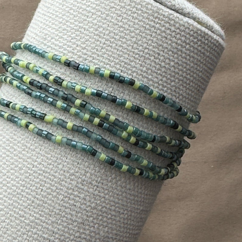 May include: A beaded wrap bracelet with multiple strands. The bracelet features small, colorful beads in shades of teal, green, yellow, and black. The bracelet is wrapped around a neutral-colored, textured surface.