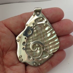 May include: A silver pendant with a pearlescent, iridescent shell inlay. The pendant has an unusual, asymmetrical shape with a decorative border and small rivet details. The shell features a swirling pattern.