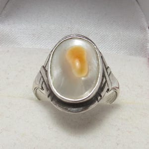 May include: A silver ring with an oval, off-white stone featuring a central, light-orange marking. The ring has a decorative band with etched details. The ring is set against a red and white background.