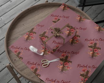 May include: A gift wrapped in mauve-colored paper with a floral cross design and the text "Rooted in Christ." The gift is tied with a brown ribbon. Scissors and tape are also visible on the table.