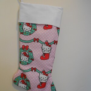 May include: A pink Christmas stocking with a white cuff and a loop for hanging. The stocking is patterned with white polka dots and features images of Hello Kitty in a Santa hat, wreaths, and red bows.