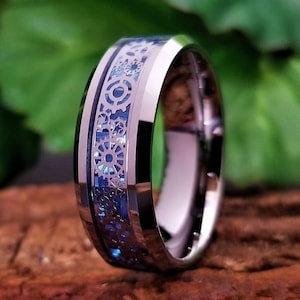 May include: A polished silver-toned metal ring featuring a central inlay of blue carbon fiber material. The inlay is overlaid with a detailed, intricate pattern of interconnected mechanical gears and cogs in a metallic silver finish.