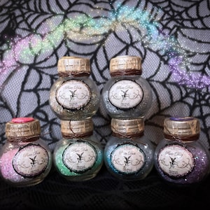 May include: Six small glass jars with cork lids and brown twine tied around the necks. Each jar contains iridescent glitter and has a label that reads "Fairy Dust". The jars are arranged in two rows of three, with the top row slightly offset from the bottom row. The jars are sitting on a black and white patterned surface.