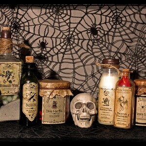 May include: A collection of eight glass bottles with cork stoppers and labels. The bottles are filled with various liquids and have labels with names like "Mermaid Wish", "Phoenix Feathers", and "The Very Last Drop". The bottles are arranged on a black surface with a spiderweb pattern background. Two white skull figurines are in front of the bottles.