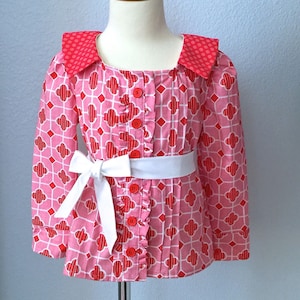 May include: A pink and red patterned button-up blouse with a white sash and a ruffled collar. The blouse is on a mannequin.