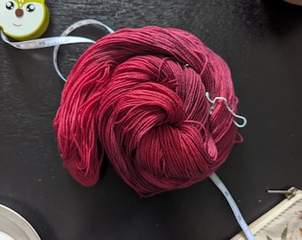 May include: A skein of red yarn with dark red and black highlights, coiled on a dark surface. A white measuring tape, a fox-shaped tape measure, a floral teacup, a silver spoon, and a small pouch are also visible.