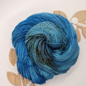 May include: A skein of blue yarn with brown and green speckles. The yarn is wound into a ball shape and is sitting on a wooden surface.