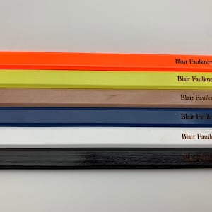 May include: Six wooden pencils in different colours: orange, yellow, brown, blue, white and black. Each pencil has the name "Blair Faulkner" engraved on it.