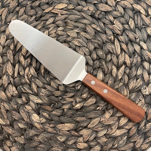 May include: A silver cake server with a wooden handle. The server is lying on a woven brown mat.