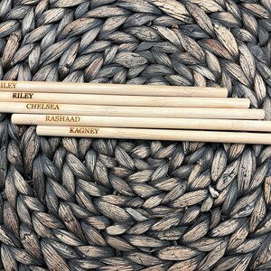 Puede incluir: Cinco palitos de bamb&uacute; natural con nombres grabados: Hailey, Riley, Chelsea, Rashaad y Kagney. Los palitos est&aacute;n dispuestos sobre una superficie de mimbre marr&oacute;n tejido.