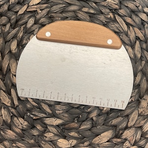 May include: A silver and wooden dough scraper with a ruler marked in centimeters from 1 to 14. The scraper is curved and has a wooden handle.