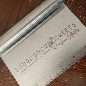 Peut inclure: Un grattoir &agrave; p&acirc;te en argent avec une r&egrave;gle en bas. Le grattoir porte la mention "SOURDOUGH SWEETS Gina Caffrider" et "BREADS, PASTA, DESSERTS & MORE!" grav&eacute;e dessus.