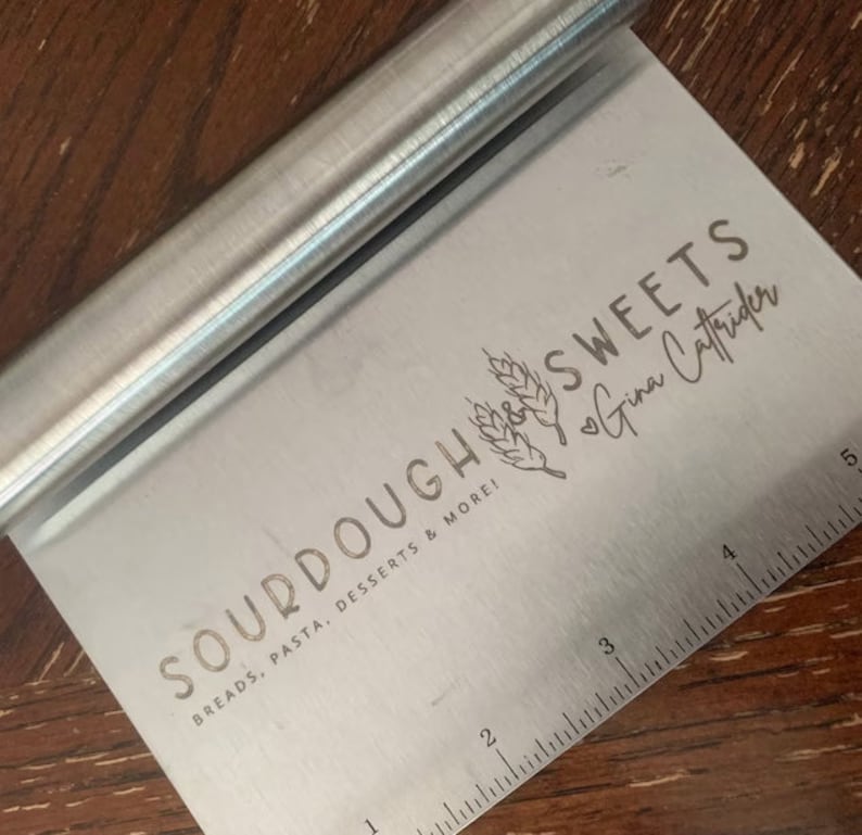 Peut inclure: Un grattoir &agrave; p&acirc;te en argent avec une r&egrave;gle en bas. Le grattoir porte la mention "SOURDOUGH SWEETS Gina Caffrider" et "BREADS, PASTA, DESSERTS & MORE!" grav&eacute;e dessus.