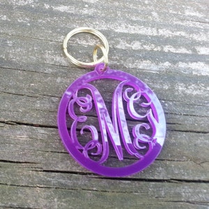 Peut inclure: Un porte-clés en acrylique violet avec un anneau de clé en or. Le porte-clés présente une initiale "M" monogramme avec des volutes décoratives.