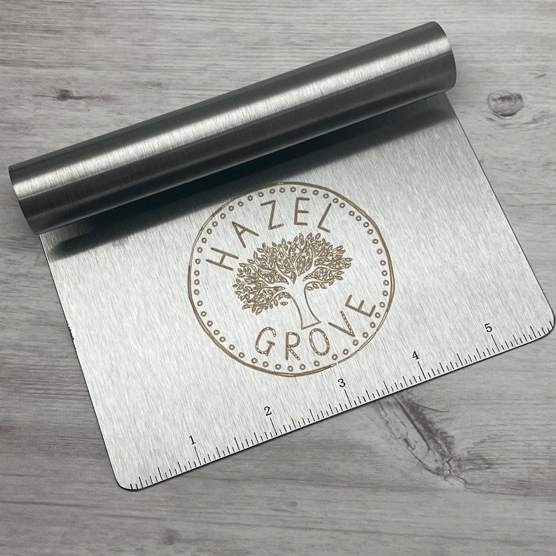 Peut inclure: Une spatule &agrave; p&acirc;tisserie en acier inoxydable avec une r&egrave;gle au bas. La spatule porte un logo grav&eacute; qui dit "Hazel Grove" avec un motif d'arbre.
