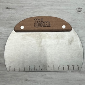 May include: A stainless steel pizza cutter with a wooden handle. The handle has the text "Yo! Zone" engraved on it. The cutter has a ruler on the bottom with measurements in centimeters from 1 to 14.