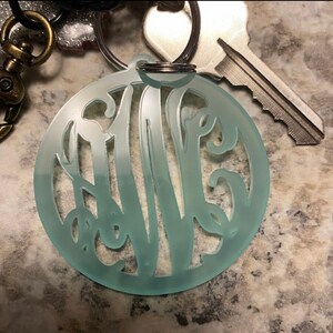 May include: A light blue acrylic keychain with a monogram design. The keychain is round and has a silver key ring.