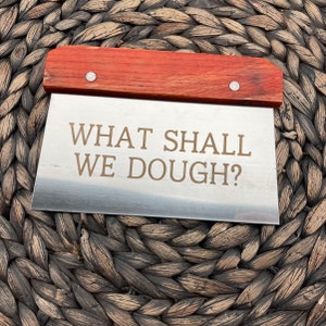 May include: A stainless steel dough scraper with a wooden handle. The scraper has the text "WHAT SHALL WE DOUGH?" engraved on it.