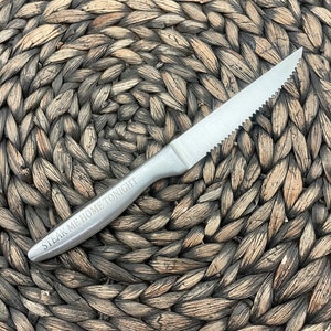 May include: A silver steak knife with a serrated edge and the words "Steak Me Home Tonight" engraved on the handle. The knife is lying on a woven brown placemat.