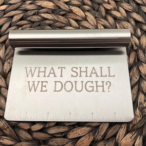 Puede incluir: Una rasqueta de masa de acero inoxidable con un mango curvo y una hoja plana y rectangular. La hoja tiene grabado el texto "WHAT SHALL WE DOUGH?" y una regla a lo largo del borde inferior. La rasqueta está sobre una superficie tejida.
