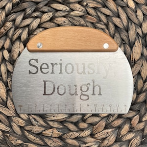 May include: A stainless steel dough scraper with a wooden handle. The scraper has a ruler on the bottom with markings in inches from 1 to 14. The scraper has the text "Seriously, Dough" engraved on the front.