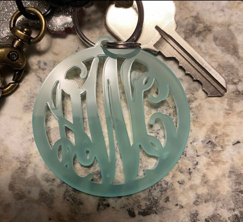 May include: A light blue acrylic keychain with a monogram design. The keychain is round and has a silver key ring.