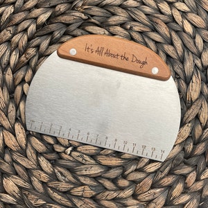 May include: A silver dough scraper with a wooden handle. The handle has the text "It's All About the Dough" engraved on it. The scraper has a ruler on the bottom edge, marked in centimeters.