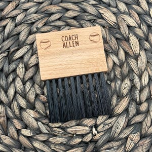 May include: A wooden brush with black bristles and a wooden handle. The handle has the text "COACH ALLEN" and two baseballs engraved on it.
