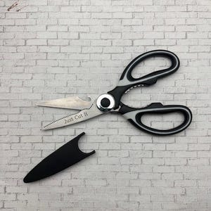 Puede incluir: Tijeras de cocina de acero inoxidable con mangos negros y blancos y un abrebotellas. El texto "Just Cut It" está grabado en la hoja. Incluye una funda protectora negra.