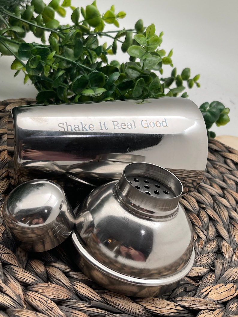 May include: A stainless steel cocktail shaker with a lid and a separate top. The shaker has the words "Shake It Real Good" engraved on the side.