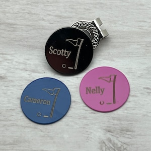 May include: Three personalised golf ball markers with a flag design. The markers are black, blue and pink. The names "Scotty", "Cameron", and "Nelly" are engraved on the markers.