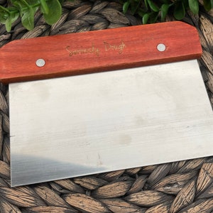 May include: A stainless steel dough scraper with a red wooden handle. The handle has the words "Seriously Dough" engraved on it.