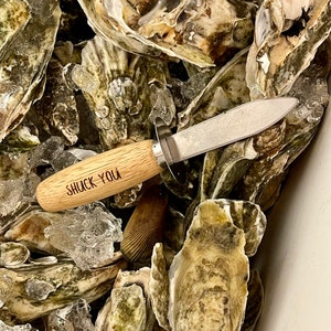 May include: A close-up of a wooden oyster shucking knife with the words "Shuck You" engraved on the handle. The knife is surrounded by a pile of fresh oysters on ice.