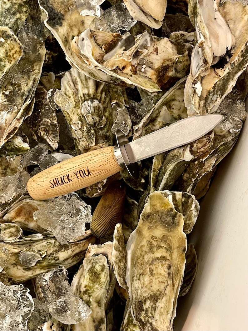 May include: A close-up of a wooden oyster shucking knife with the words "Shuck You" engraved on the handle. The knife is surrounded by a pile of fresh oysters on ice.