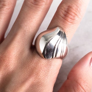 Könnte beinhalten: Ein silberfarbener Ring mit einer abgerundeten, bauchigen Form und vertikalen Rillen. Der Ring wird an einem Finger getragen und zeigt seine polierte Oberfläche und sein einzigartiges Design. Der Schmuck befindet sich vor einem neutralen Hintergrund.