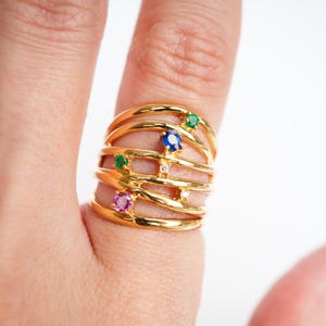 Puede incluir: Anillo de oro con múltiples bandas y piedras preciosas de colores. El anillo presenta una piedra rosa, una azul y dos verdes.