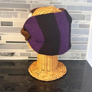 handmade crochet headband in shades of purple, black, brown, and white. The headband is displayed on a wicker head form. The design features vertical stripes of black and purple, with brown and white accents.