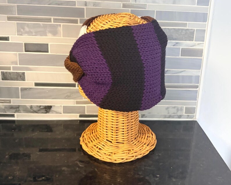 handmade crochet headband in shades of purple, black, brown, and white. The headband is displayed on a wicker head form. The design features vertical stripes of black and purple, with brown and white accents.