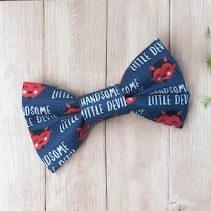 May include: A navy blue bow tie with a repeating pattern of red devil faces and the text "LITTLE DEVIL" and "HANDSOME SOME". The bow tie is made of fabric and is suitable for special occasions.